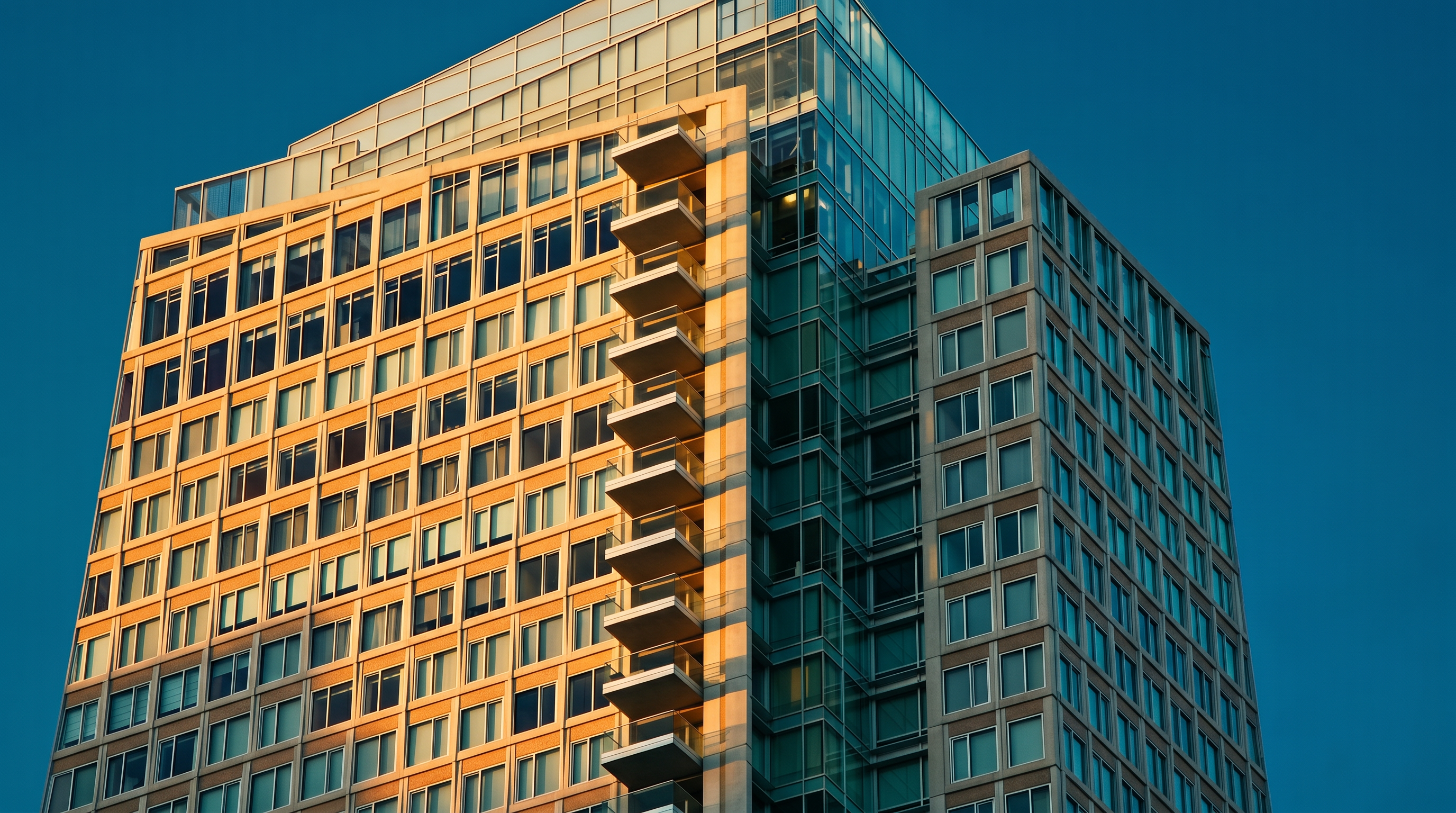 This image captures a low-angle, cinematic perspective of a modern high-rise residential building against a clear, deep blue sky. The structure features a mix of glass curtain walls and a grid of windows, with a prominent vertical stack of balconies extending along the side. The warm, golden light of late afternoon highlights the architectural geometry and the contrast between the reflective glass and the building's facade.