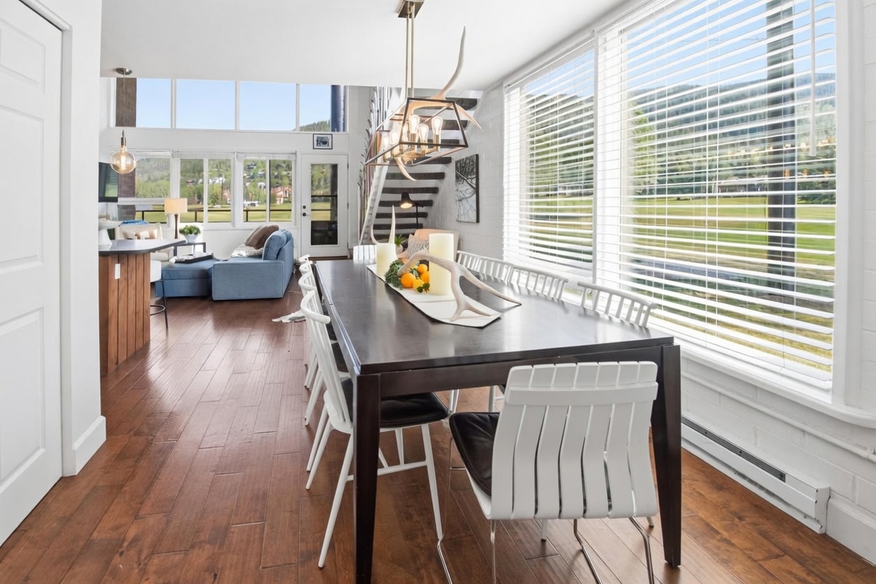 This bright dining area features a dark wood table surrounded by modern white chairs, set against a wall of large windows with horizontal blinds that offer a view of the outdoors. An antler-inspired chandelier hangs above the table, which is decorated with a centerpiece of candles and fruit. The open-concept space flows seamlessly into a living area with a blue sofa, all set upon warm-toned wood flooring.