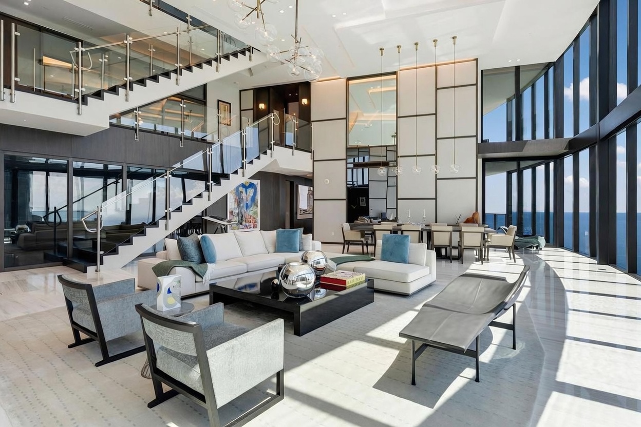 This expansive, double-height living room features a sophisticated open-concept design with a striking floating staircase and a wall of floor-to-ceiling windows overlooking the ocean. The space is furnished with a large sectional sofa, contemporary armchairs, and a central coffee table, all set against a backdrop of neutral tones and high-end finishes. The room is bathed in natural light, creating an airy and luxurious atmosphere that seamlessly connects the indoor living area to the dining space in the background.