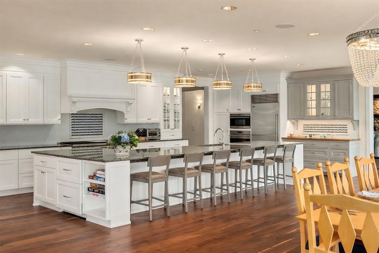 This spacious, high-end kitchen features a large central island with seating for five, topped with a dark stone surface and illuminated by four matching pendant lights. The room is anchored by white cabinetry, a custom range hood, and a separate grey-toned cabinet section, all set against rich dark wood flooring. The perspective captures the expansive layout, highlighting the seamless transition between the functional cooking area and the adjacent dining space.