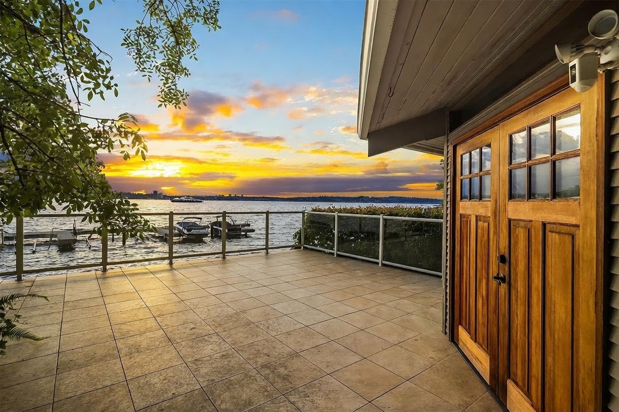 This expansive, tiled deck offers a stunning waterfront view at sunset, featuring a glass-paneled railing that maintains an unobstructed perspective of the lake and distant city skyline. To the right, a set of double wooden doors with glass panes provides access to the home, while a security camera is mounted above the frame. The scene captures a serene, high-end outdoor living space perfect for relaxation or entertaining against a vibrant, colorful sky.