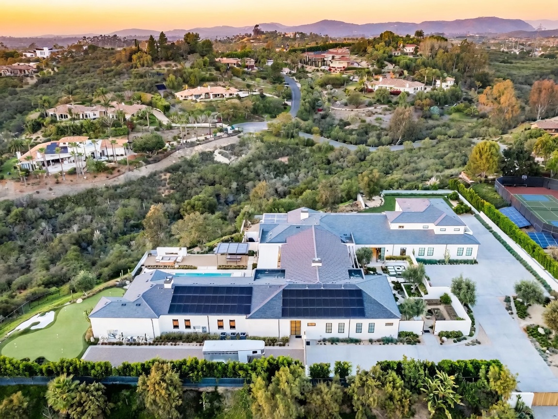 This high-angle aerial view captures a sprawling, modern estate nestled in a hilly landscape at sunset. The property features a large white residence with dark roofing, solar panels, a private swimming pool, and a putting green, all surrounded by lush greenery and rolling hills. A tennis court is visible on the right side of the property, emphasizing the expansive and luxurious nature of the grounds.