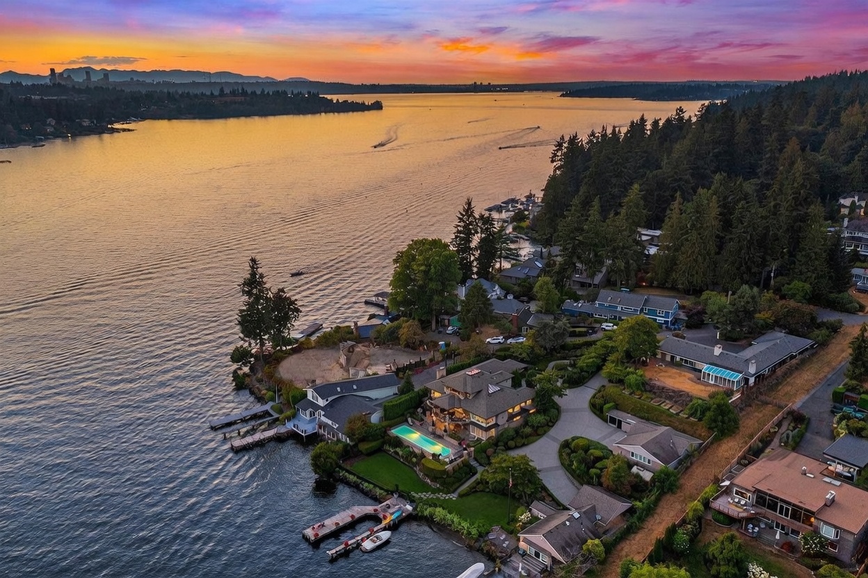 This stunning aerial perspective captures a luxury waterfront estate at sunset, showcasing its prime location on a serene lake. The property features a multi-level residence with a private swimming pool, a manicured lawn, and a dedicated boat dock extending into the water. The surrounding landscape is lush with mature trees, and the golden hour light creates a dramatic, cinematic atmosphere over the expansive water view.