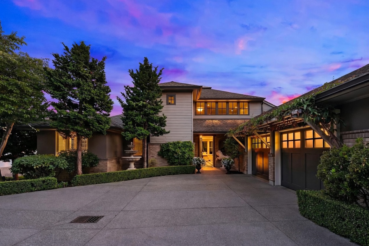 This front-facing view captures a multi-story residence at dusk, featuring a concrete driveway leading to a multi-car garage with a decorative wooden trellis overhead. The home showcases a blend of horizontal siding and brick accents, with warm interior lighting glowing through large windows and the main entryway. A manicured hedge and a stone fountain add a touch of elegance to the landscaped front yard, all set against a vibrant, colorful sunset sky.