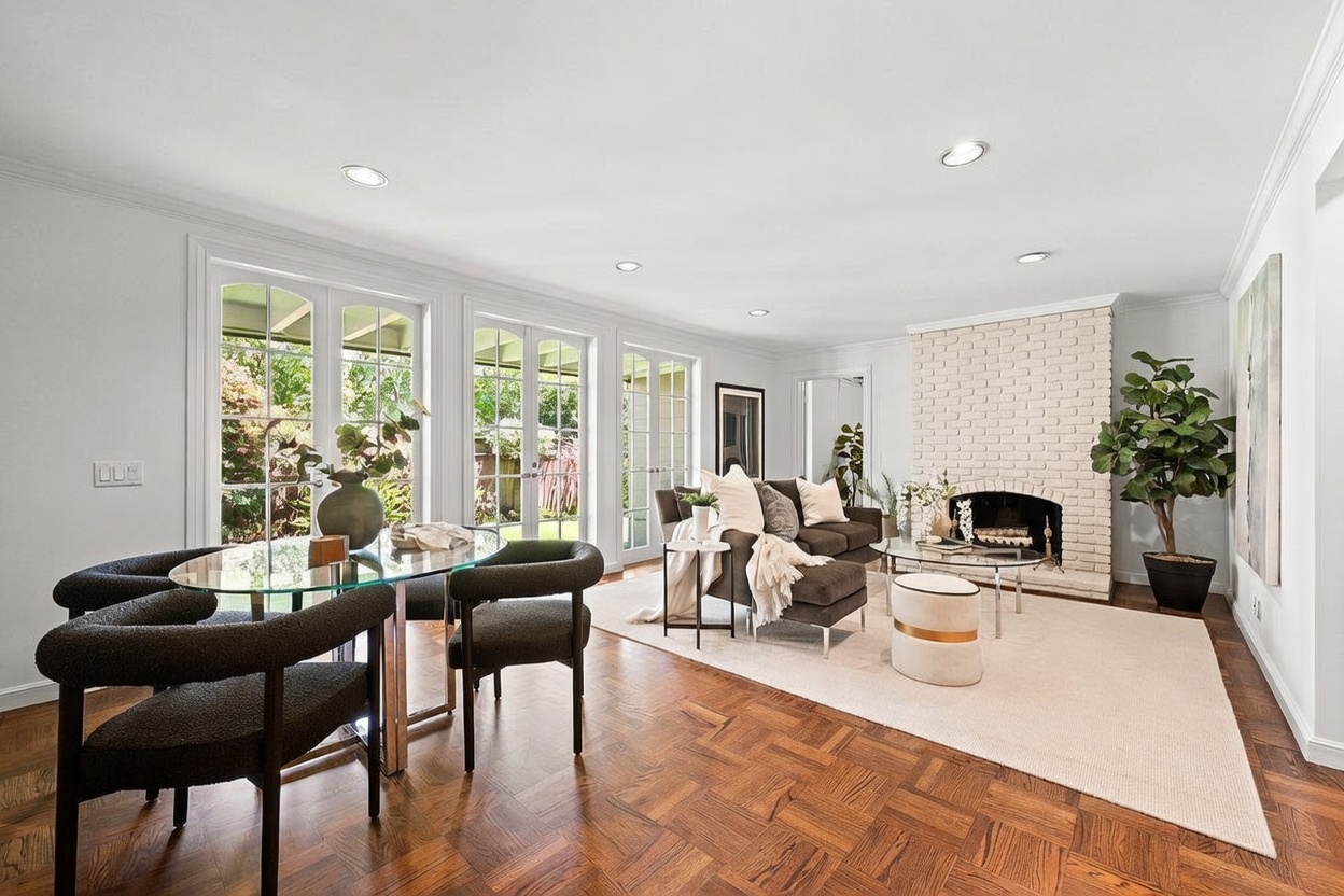 This open-concept living and dining area features rich, patterned wood flooring and a bright, neutral color palette. A prominent white brick fireplace serves as a focal point, complemented by a comfortable seating arrangement and a glass-topped dining table. Large glass doors provide ample natural light and a seamless connection to the outdoor space, creating an inviting and airy atmosphere.