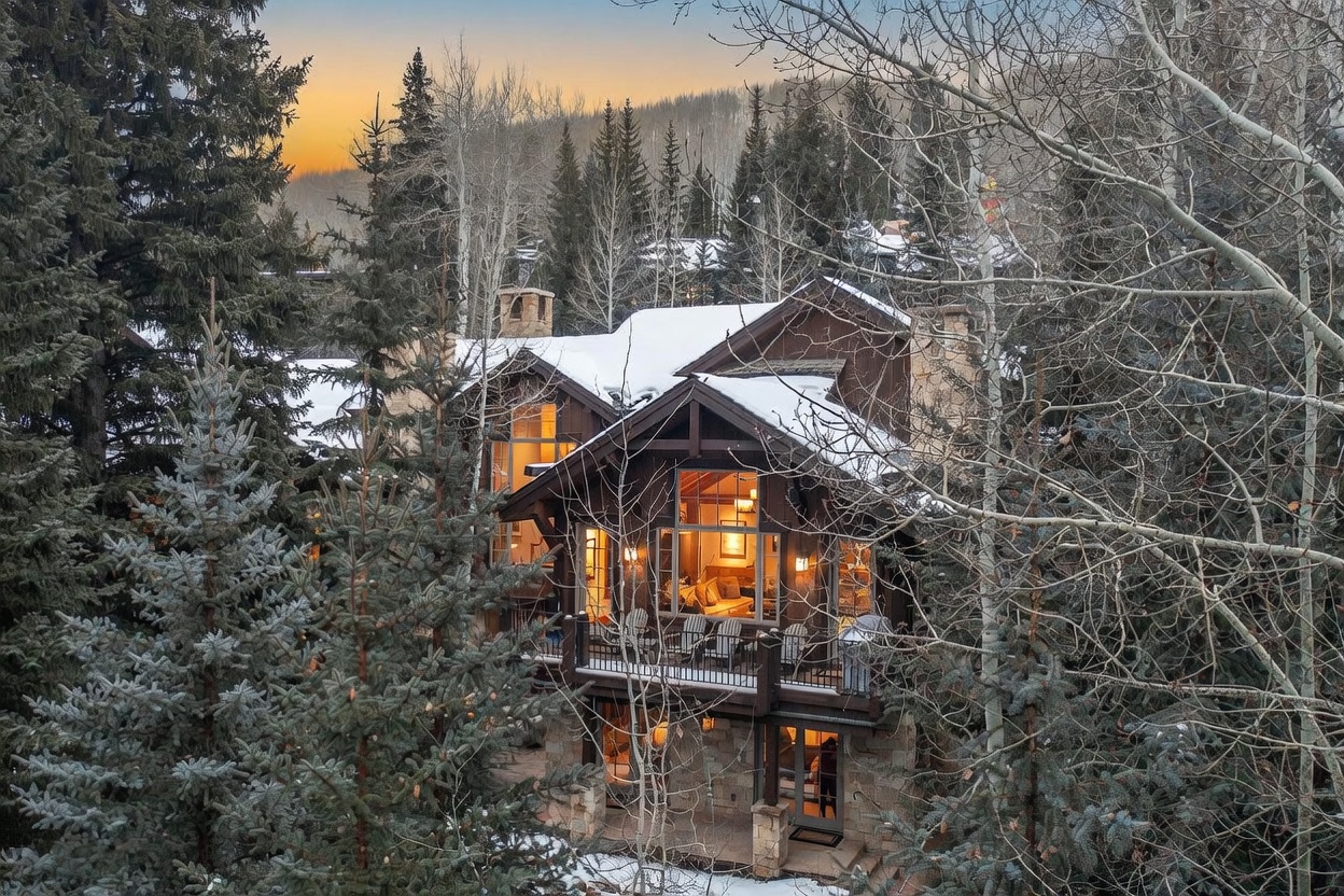 This mountain-style residence is captured from an elevated perspective, nestled within a dense forest of evergreen and aspen trees. The home features a multi-level design with a prominent wooden deck, stone foundation, and large windows that reveal a warm, inviting interior glow. The scene is set during twilight, highlighting the contrast between the dark, snow-dusted architecture and the soft, golden light emanating from within.
