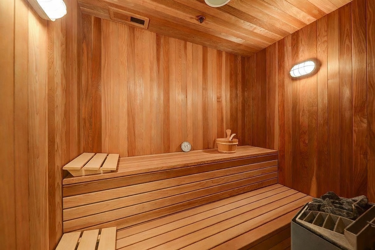 This image showcases a dedicated sauna room finished in warm, light-toned wood paneling throughout the walls and tiered benches. The space includes a built-in heater with stones in the foreground, a wooden bucket, and a small wall-mounted clock, creating a serene and functional wellness area. The perspective is a wide-angle interior shot that captures the full depth of the wood-lined enclosure and its integrated lighting fixtures.