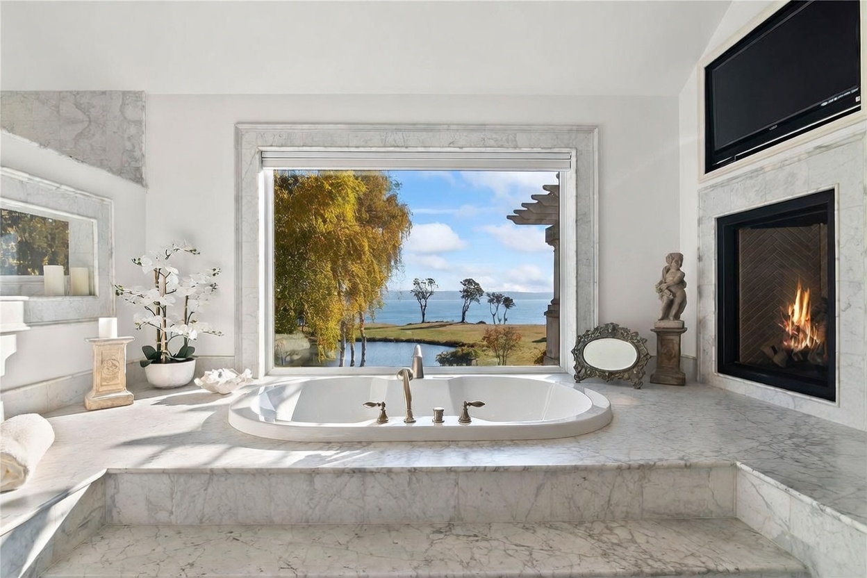 This luxurious primary bathroom features a central soaking tub set into a stone-clad platform, perfectly positioned in front of a large window that frames a scenic waterfront view. To the right, a fireplace with a stone surround adds warmth, while decorative accents like a small statue and a mirror enhance the elegant aesthetic. The room is finished with light-toned stone surfaces, creating a serene and sophisticated atmosphere.