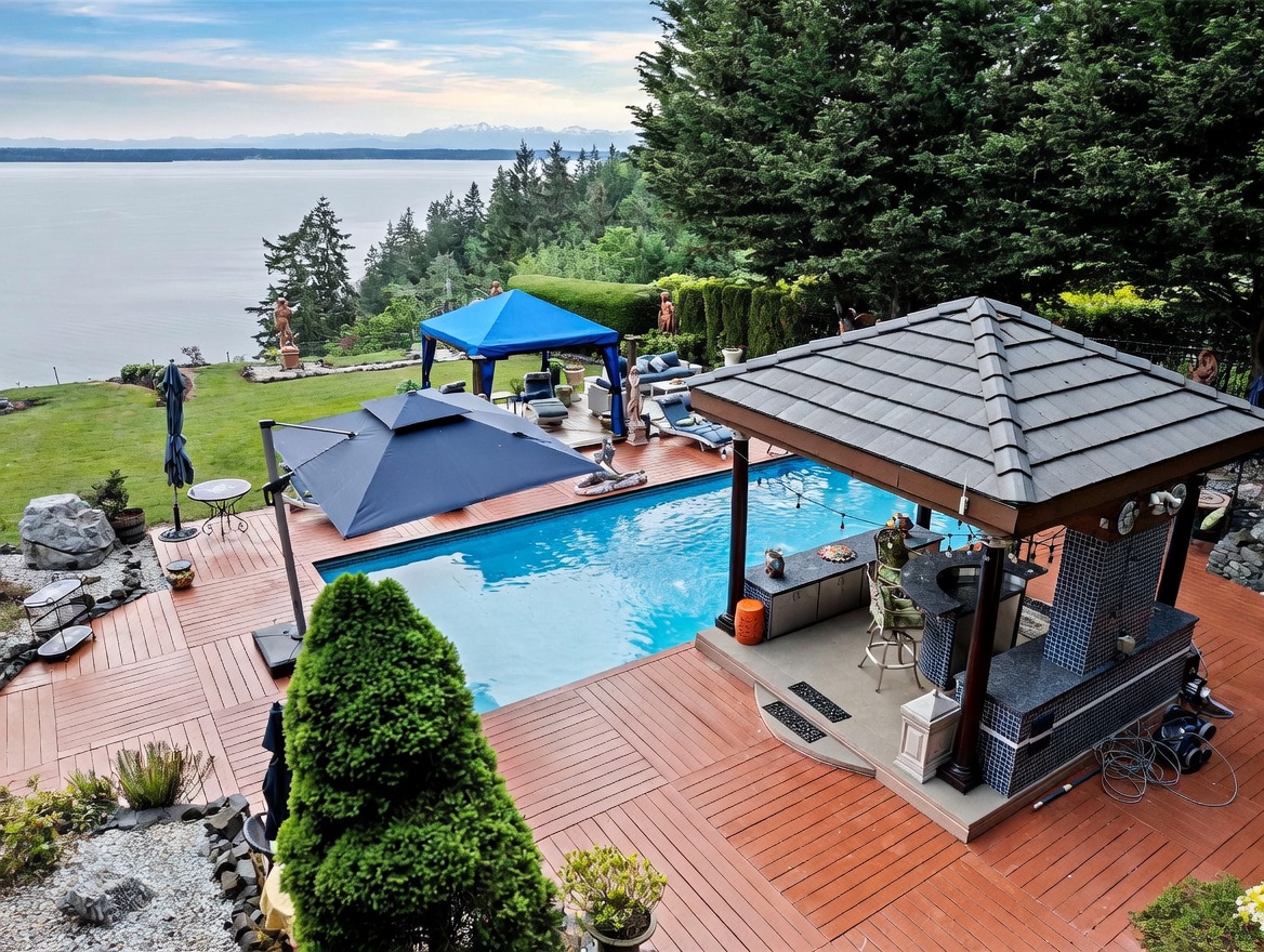 This high-angle aerial view captures a luxurious outdoor living space featuring a rectangular swimming pool set into a spacious wooden deck. The area includes a covered outdoor kitchen and bar structure, a large blue canopy, and a cantilevered umbrella, all overlooking a scenic waterfront with distant mountains. The expansive lawn and mature trees create a serene, resort-like atmosphere perfect for entertaining.