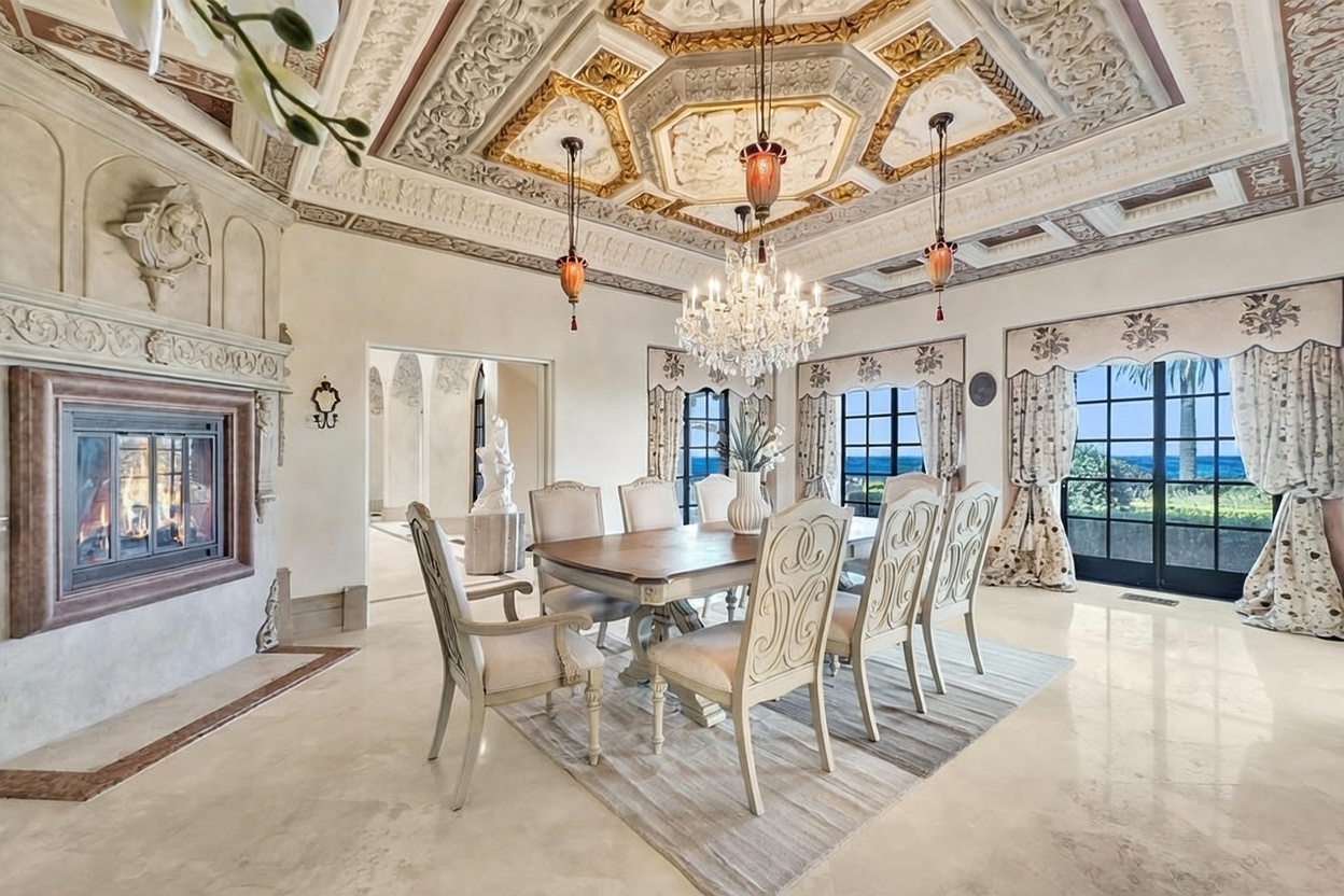 This elegant dining room features a formal table set for six, positioned beneath an ornate, coffered ceiling with intricate molding and a crystal chandelier. To the left, a stone fireplace with a decorative mantel serves as a focal point, while large windows with patterned drapery offer a view of the ocean. The room is finished with polished stone flooring and a neutral color palette, creating a sophisticated and luxurious atmosphere.