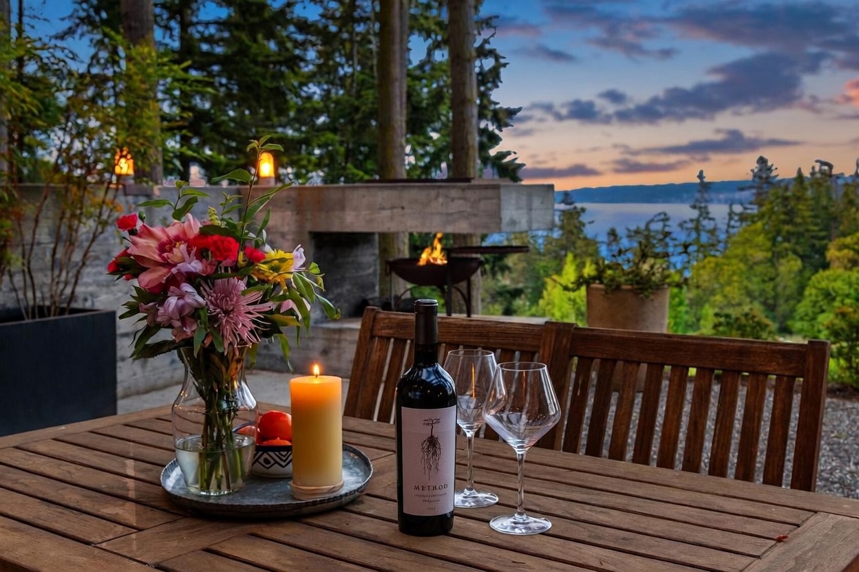 This inviting outdoor dining space features a wooden table set with a bouquet of flowers, a lit candle, and a bottle of wine, creating a warm and intimate atmosphere. In the background, a concrete outdoor fireplace with a burning fire provides a cozy focal point, while the expansive view of the water and forest at sunset adds a serene, high-end feel. The perspective is captured from a seated eye-level, emphasizing the tranquil lifestyle and scenic beauty of the property.