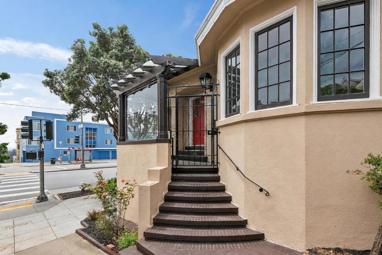 This exterior view captures the front entryway of a home, featuring a set of dark brick steps leading up to a secure metal gate and a vibrant red front door. The home is finished in a warm-toned stucco and includes a small, glass-enclosed porch area with a decorative pergola-style roof. The perspective is from the sidewalk, showing the relationship between the home's entrance and the adjacent street corner.