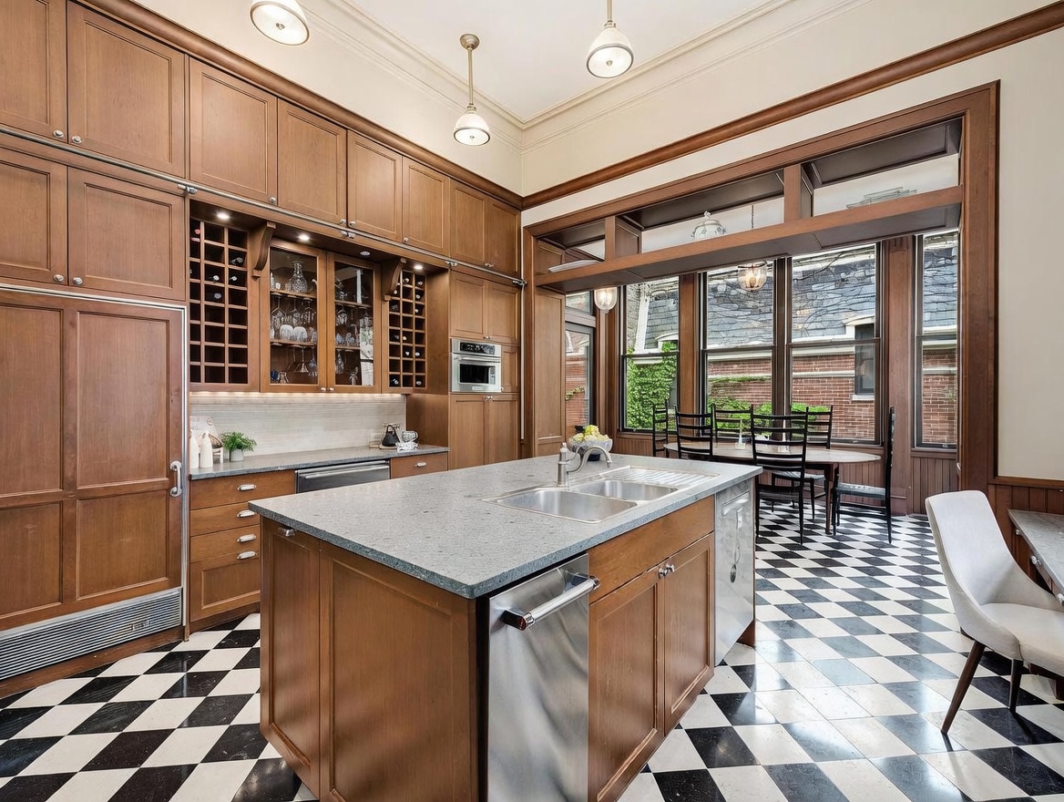 This spacious kitchen features warm-toned wood cabinetry, a large central island with a stone countertop, and a striking black-and-white checkered floor. The space is illuminated by multiple pendant lights and large windows that provide a view of the outdoor brick wall and greenery. A dining area is visible in the background, creating an open and inviting atmosphere suitable for both cooking and entertaining.