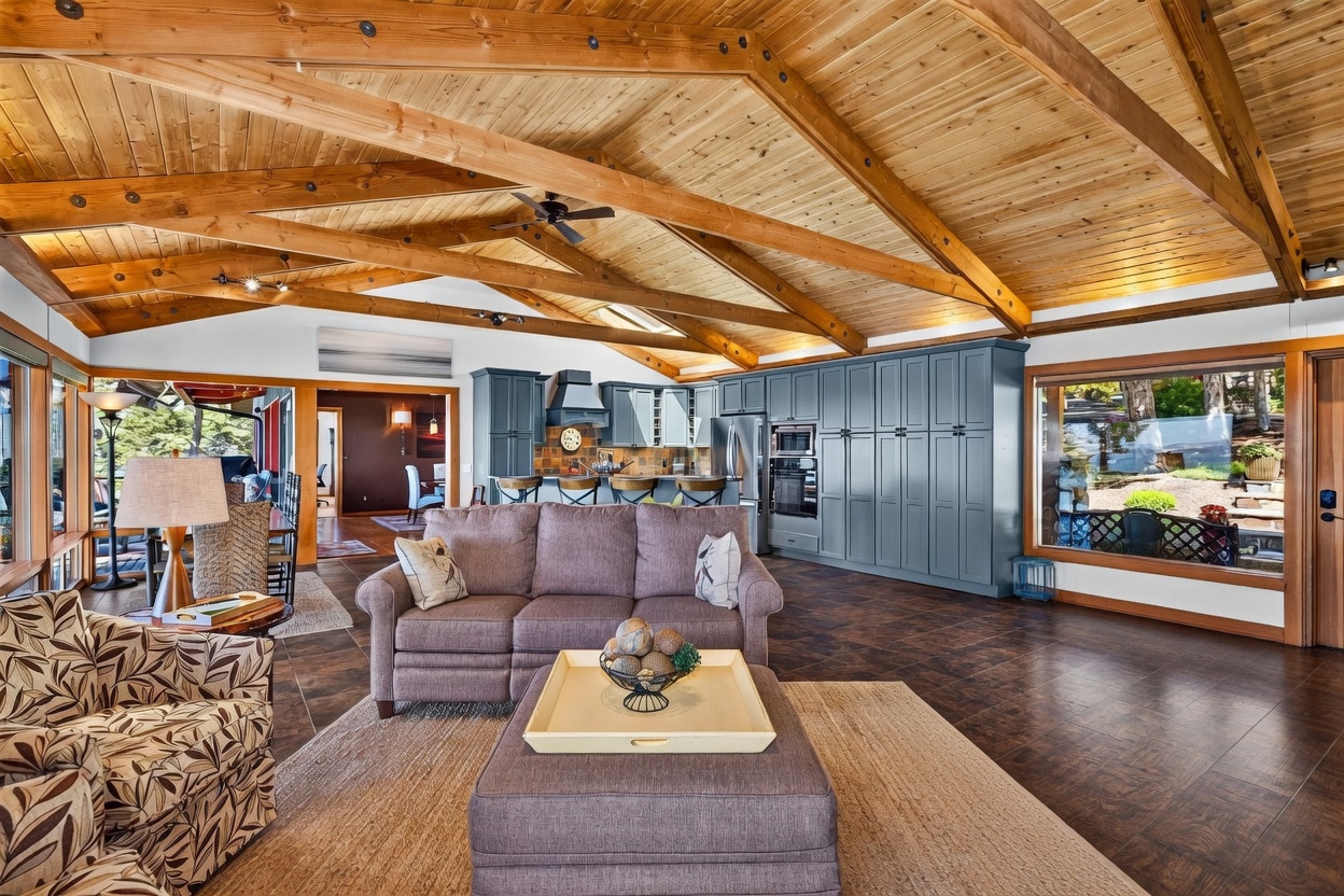 This spacious open-concept living area features a dramatic vaulted ceiling with exposed wooden beams and a warm wood-paneled finish. The room is anchored by a comfortable seating arrangement with two sofas and a central ottoman, while a kitchen with blue cabinetry and a breakfast bar is visible in the background. Large windows provide ample natural light and views of the surrounding greenery, creating an inviting and airy atmosphere.