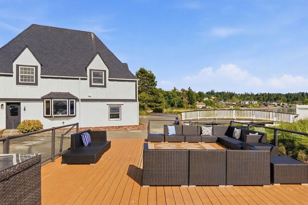 This spacious outdoor deck features a large, modular dark wicker sectional sofa arranged in a U-shape, providing ample seating for entertaining. The deck is constructed with light-toned composite planks and is enclosed by a modern glass railing that offers an unobstructed view of a white pedestrian bridge and the surrounding landscape. The scene is captured from a slightly elevated perspective, highlighting the seamless transition between the home's exterior and the scenic waterfront setting.