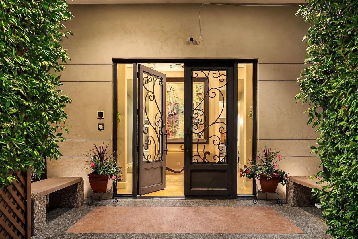 This elegant entryway features a grand double-door entrance with intricate wrought-iron scrollwork and glass panels. The space is framed by lush greenery on both sides, with stone benches and potted plants creating a welcoming, symmetrical aesthetic. A security camera is mounted above the doorway, and the warm lighting highlights the textured wall finish and tiled landing.