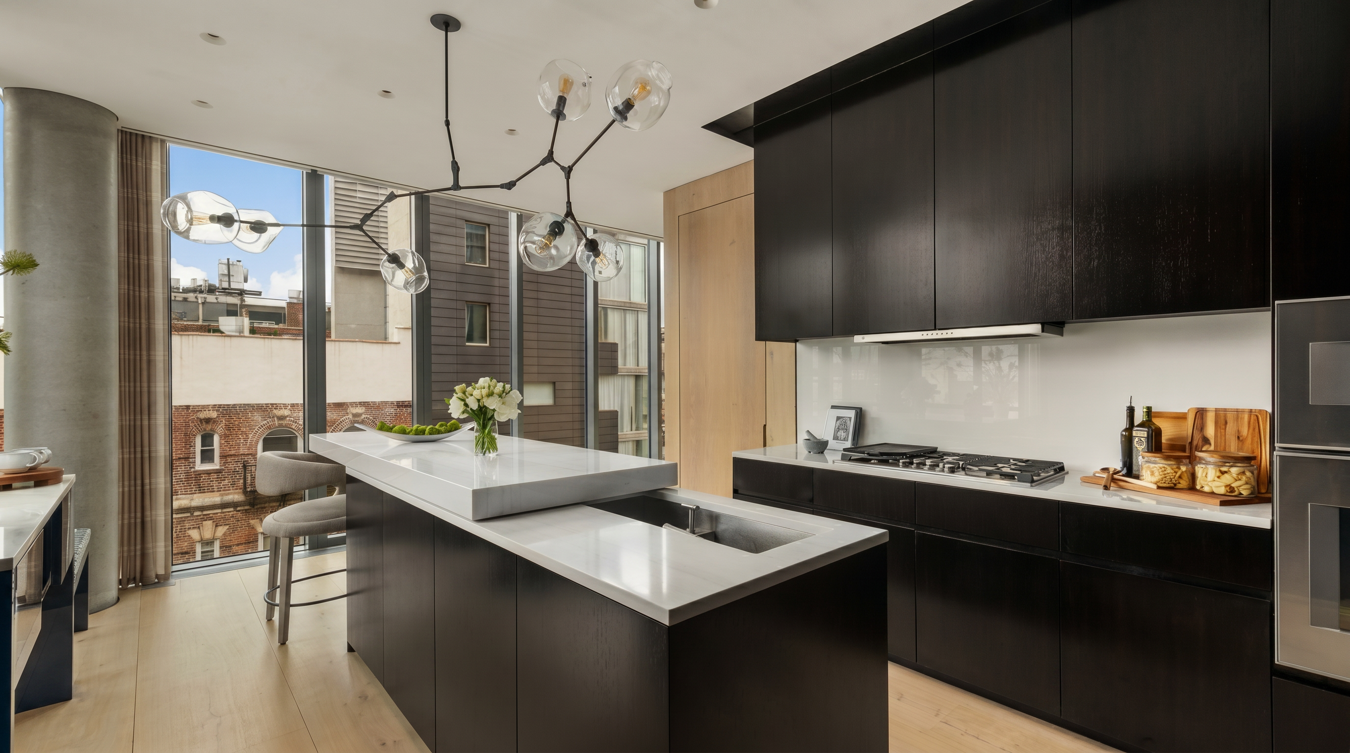 This modern, high-end kitchen features sleek dark cabinetry contrasted by light wood flooring and a large central island with a white stone countertop. A unique, sculptural chandelier hangs above the island, while large windows provide ample natural light and an urban view. The space is designed with a minimalist aesthetic, incorporating integrated appliances and a clean, functional layout.