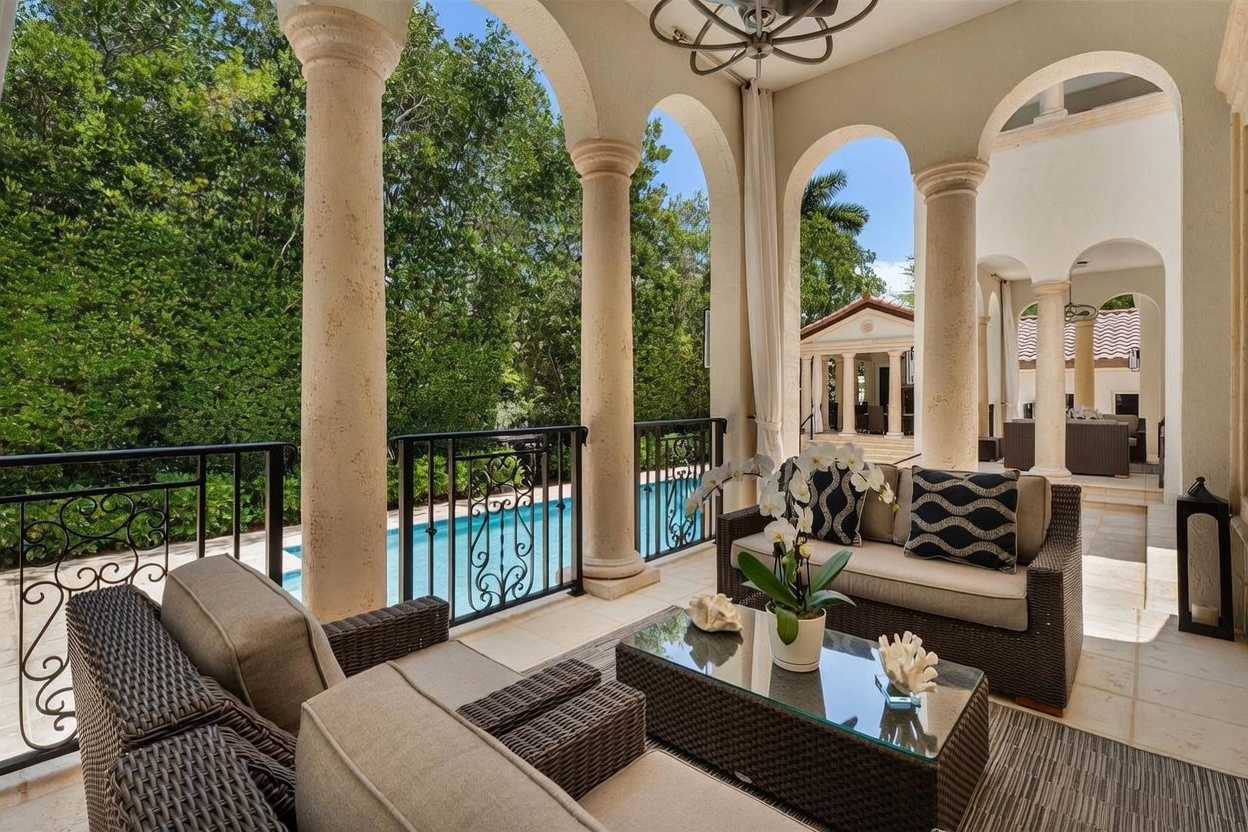 This elegant covered patio features classic architectural columns and a wrought-iron railing overlooking a private swimming pool. The space is furnished with comfortable outdoor seating, including a sofa and armchairs arranged around a glass-topped coffee table decorated with white coral accents. The view extends to a lush, tree-lined backyard and a secondary pool house structure, creating a serene and luxurious outdoor living environment.