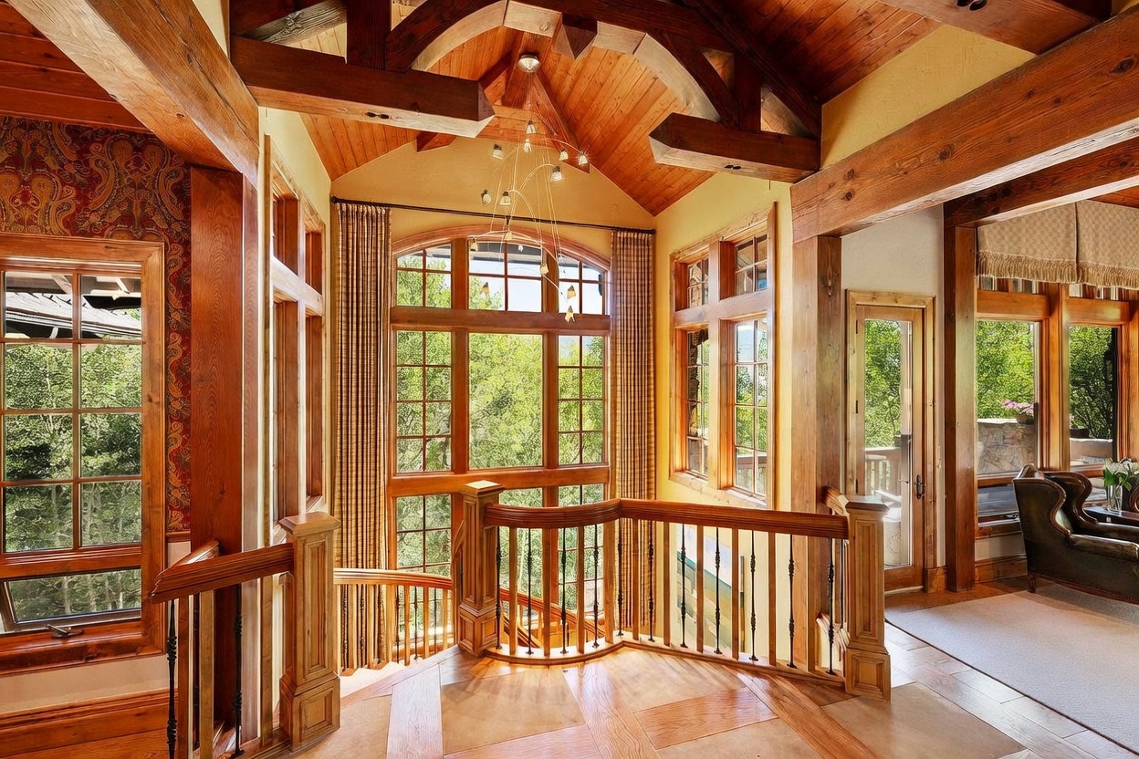 This elegant landing area features a grand staircase with a wooden railing and iron balusters, set against a backdrop of tall, multi-paned windows that flood the space with natural light. The room is defined by its rich wood-paneled vaulted ceiling with exposed timber beams and a sophisticated chandelier. To the left, patterned wallpaper adds a touch of traditional charm, while the open layout provides a seamless transition into the adjacent living area.