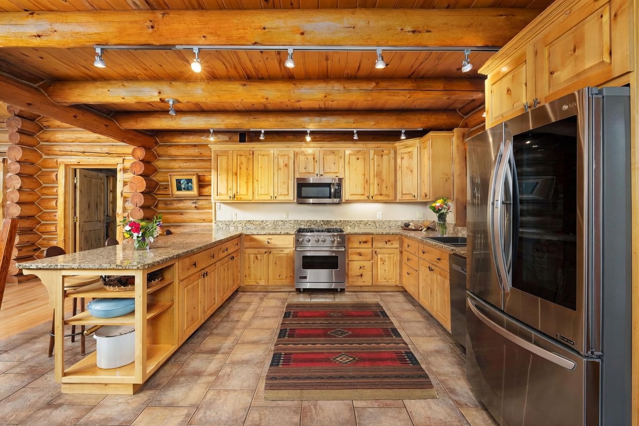 This rustic kitchen features light wood cabinetry, granite countertops, and stainless steel appliances, including a professional-style range and a large refrigerator. The space is defined by exposed log walls and a beamed ceiling, creating a warm, cabin-inspired atmosphere. A central island with open shelving and a patterned area rug on the tiled floor complete the functional and inviting layout.