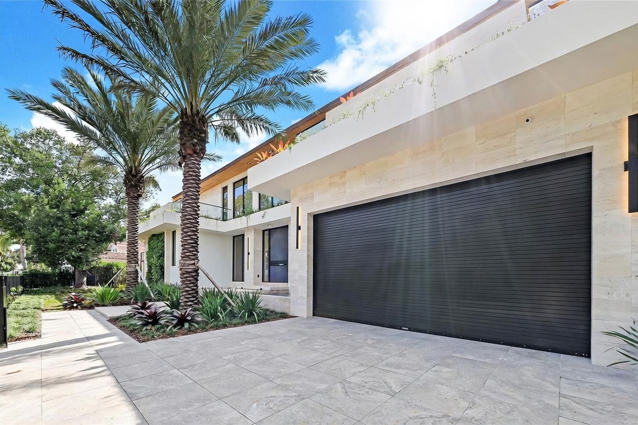 This modern, multi-level residence features a striking facade with light-toned stone cladding and a prominent, dark-finished garage door. A tall palm tree stands in the foreground, complemented by manicured landscaping and a paved driveway that leads to the main entrance. The architectural design emphasizes clean lines and a contemporary aesthetic, creating a sophisticated and welcoming curb appeal.