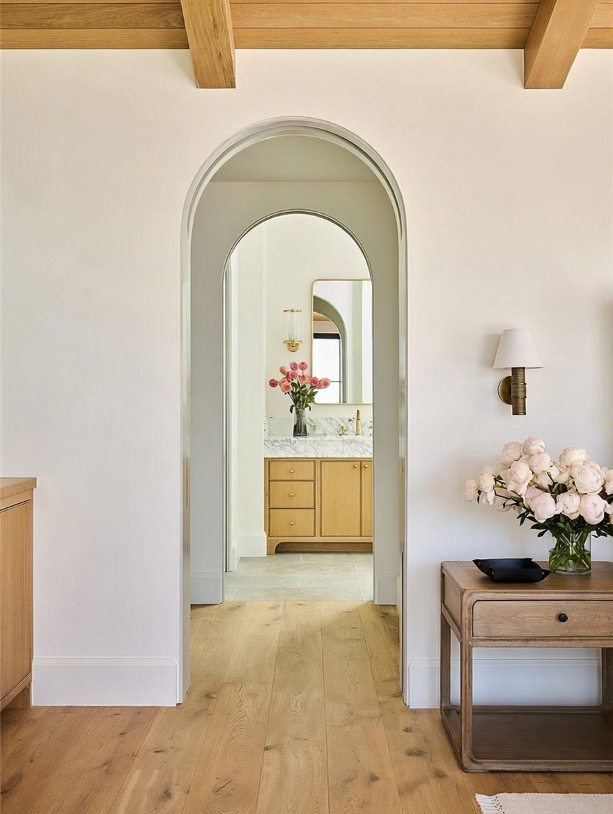 This view captures a transition space featuring a prominent arched doorway that leads into a bright bathroom. The foreground includes a light wood side table topped with a large bouquet of pink flowers, complemented by a wall-mounted sconce. The warm wood flooring and exposed ceiling beams create a cohesive, inviting atmosphere throughout the home.
