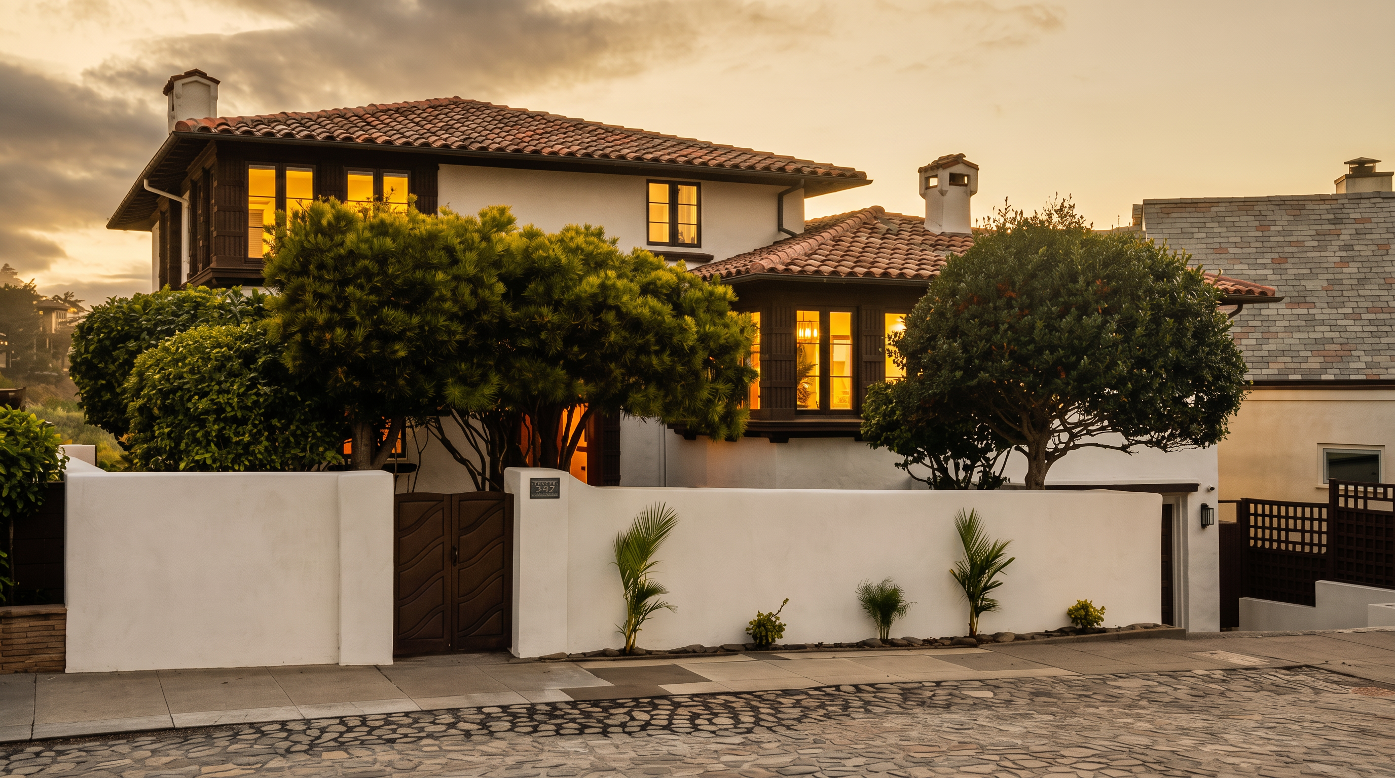 This Mediterranean-style home features a classic white stucco facade with dark wood trim and a traditional red tile roof. A solid white perimeter wall with a gated entrance provides privacy, while mature trees and small landscaping accents frame the front of the property. The image is captured at dusk, highlighting the warm glow from the interior windows against the deep blue twilight sky.