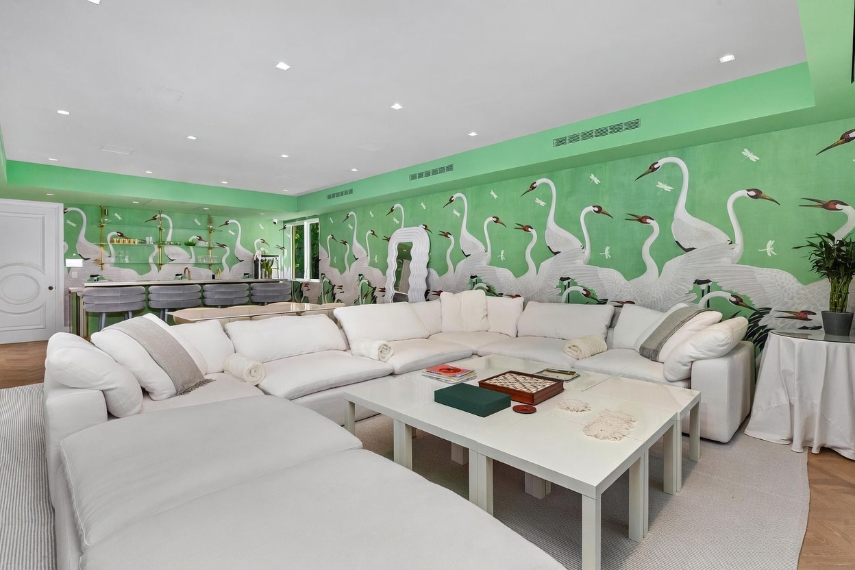 This spacious living room features a large, plush white sectional sofa arranged around a central coffee table, creating a comfortable seating area. The room is defined by a striking, vibrant green wall mural depicting a flock of cranes, which serves as a bold focal point. In the background, a built-in bar area with glass shelving and bar stools adds a sophisticated touch, while recessed lighting illuminates the entire space.