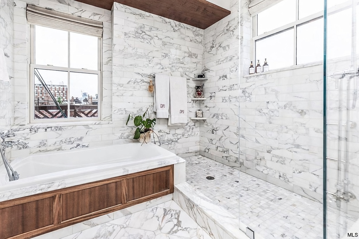 This luxurious primary bathroom features a wet room design with a deep soaking tub positioned next to a spacious walk-in shower. The space is finished with light-toned stone wall tiles and a mosaic tile floor, complemented by a warm wood-toned vanity base and ceiling accent. A large window provides natural light, creating an airy and sophisticated atmosphere.