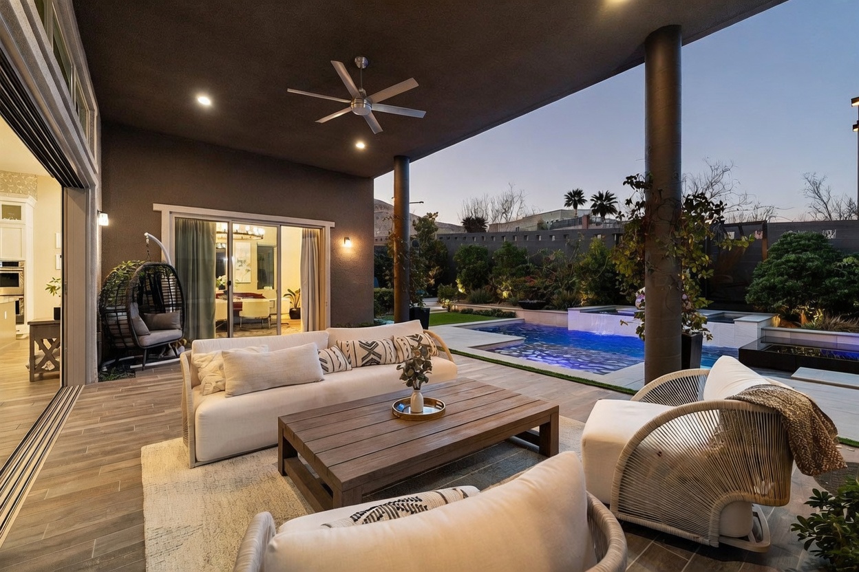 This luxurious covered patio offers a seamless transition between indoor and outdoor living, featuring a modern sofa and armchairs arranged around a low wooden coffee table. The space is illuminated by recessed lighting and a ceiling fan, overlooking a pristine swimming pool and landscaped backyard at dusk. Large sliding glass doors open to reveal a glimpse of the interior dining area, creating an inviting and sophisticated atmosphere for entertaining.