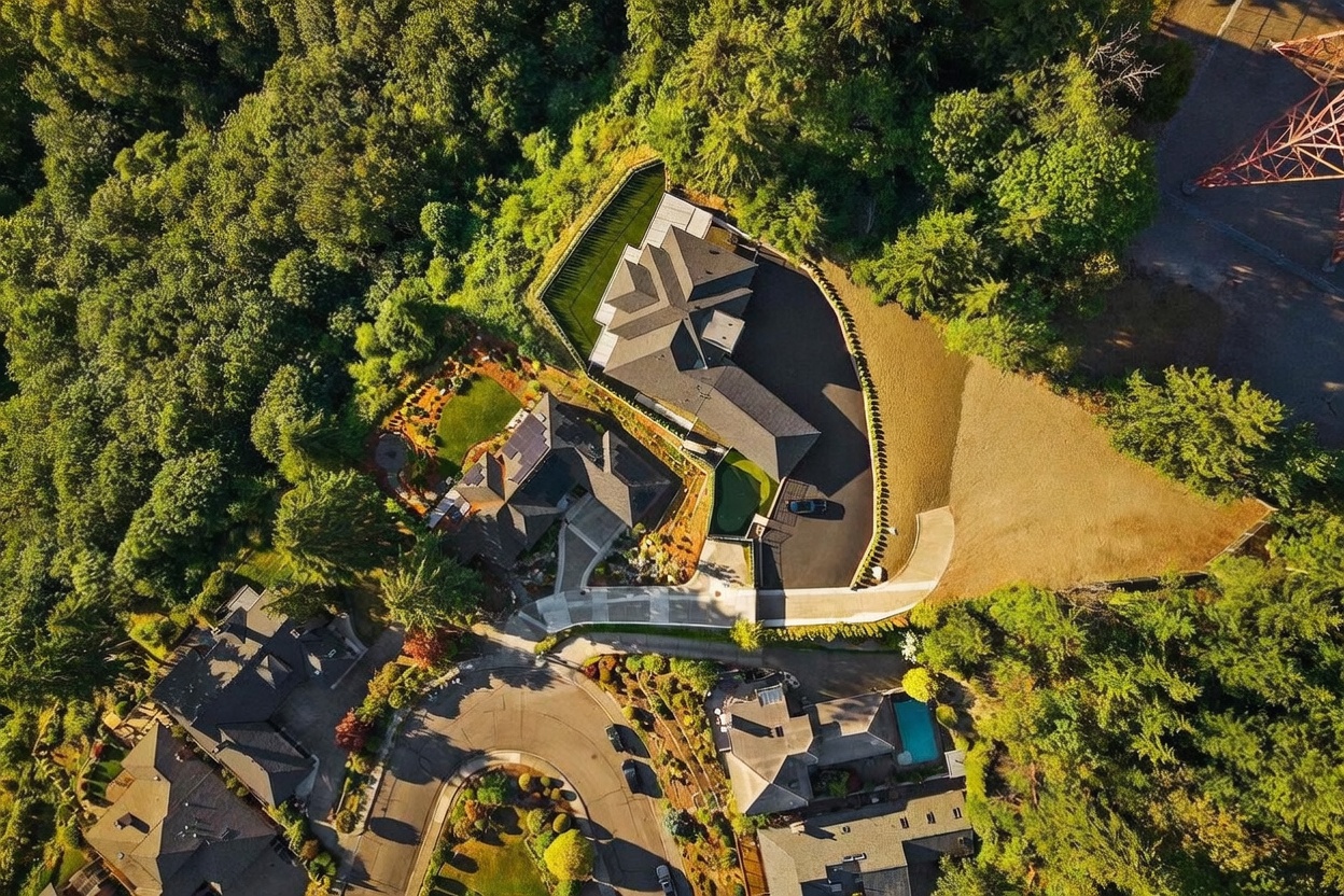This high-angle aerial view captures a sprawling residential property nestled within a lush, wooded landscape. The image highlights the complex rooflines of the main residence, a large paved driveway, and a manicured lawn area, all set against a backdrop of dense forest. A nearby power transmission tower is visible in the upper right corner, providing a sense of scale and location for this secluded estate.