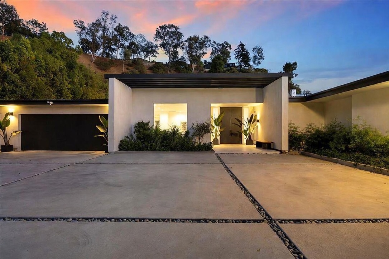 This modern, single-story residence features a minimalist architectural design with a flat roofline and a clean, light-colored facade. A wide concrete driveway with integrated stone-filled expansion joints leads to a prominent entryway and an attached garage, framed by lush landscaping and potted plants. The scene is captured at dusk, with warm interior lighting glowing through the windows and exterior accent lights highlighting the structure against a vibrant sunset sky.
