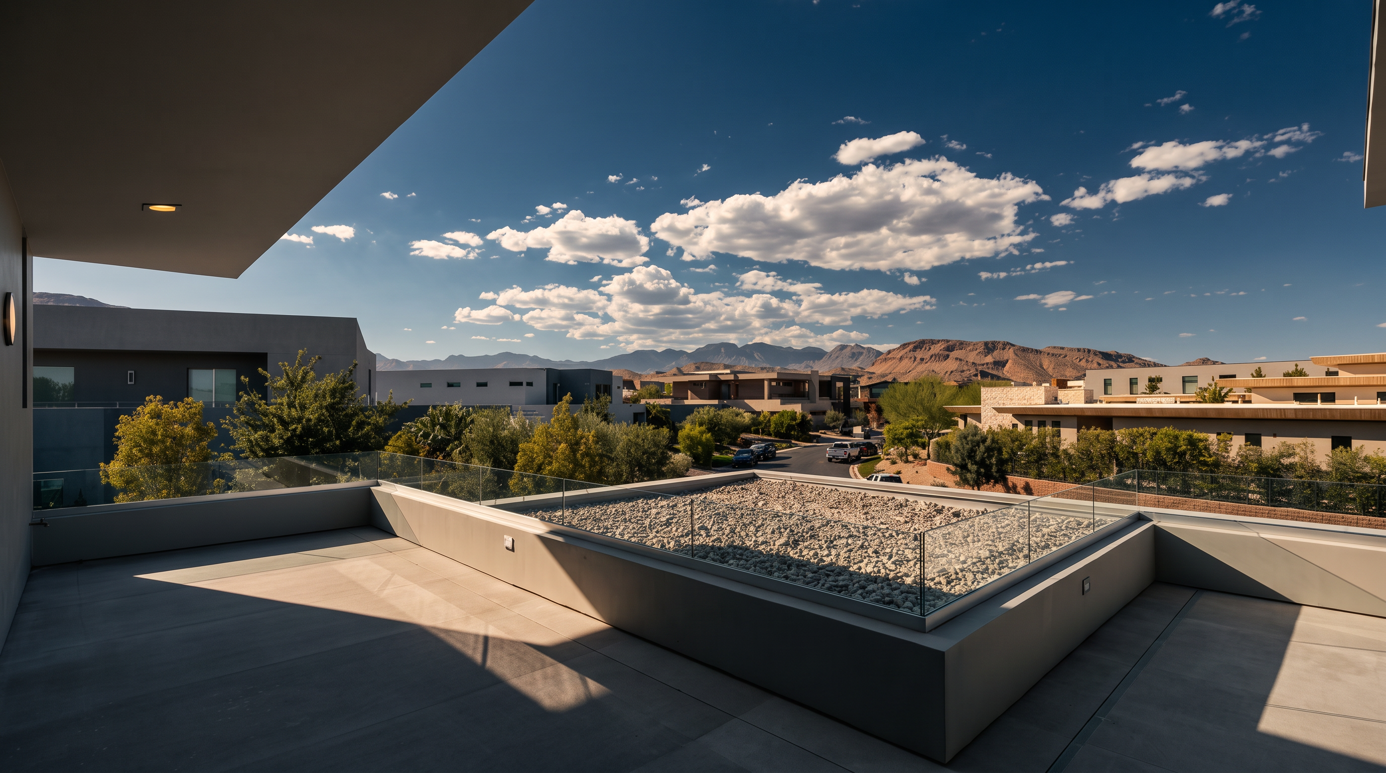 This modern, elevated balcony offers a spacious outdoor living area with contemporary lounge seating and a glass railing that provides unobstructed views of the surrounding neighborhood and distant mountains. A central, square-shaped feature filled with decorative stones adds a unique architectural element to the deck. The bright, sunny atmosphere and clear blue sky create an inviting and luxurious outdoor space.