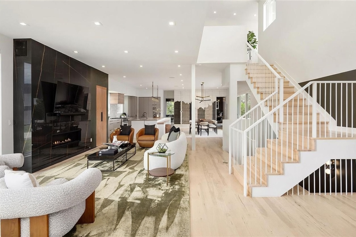 This open-concept living area features a striking dark, reflective wall panel that serves as a focal point for the television and fireplace. The space is furnished with a curved white sofa and two leather armchairs, all arranged on a large patterned rug over light wood flooring. A staircase with white railings leads to the upper level, while the background reveals a bright, modern kitchen and dining area, creating a cohesive and airy atmosphere.