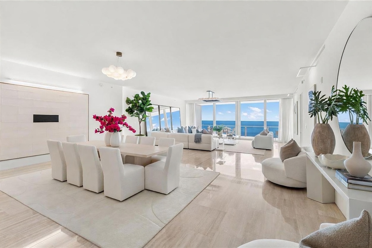 This expansive, open-concept living and dining area features a bright, minimalist aesthetic with polished light-toned flooring and crisp white walls. The space is anchored by a large dining table with eight upholstered chairs and a spacious seating area centered around a plush white sofa, all oriented toward expansive windows that provide a panoramic view of the ocean. The room is accented by a tall potted tree, vibrant floral arrangements, and a large circular mirror, creating a sophisticated and airy atmosphere.
