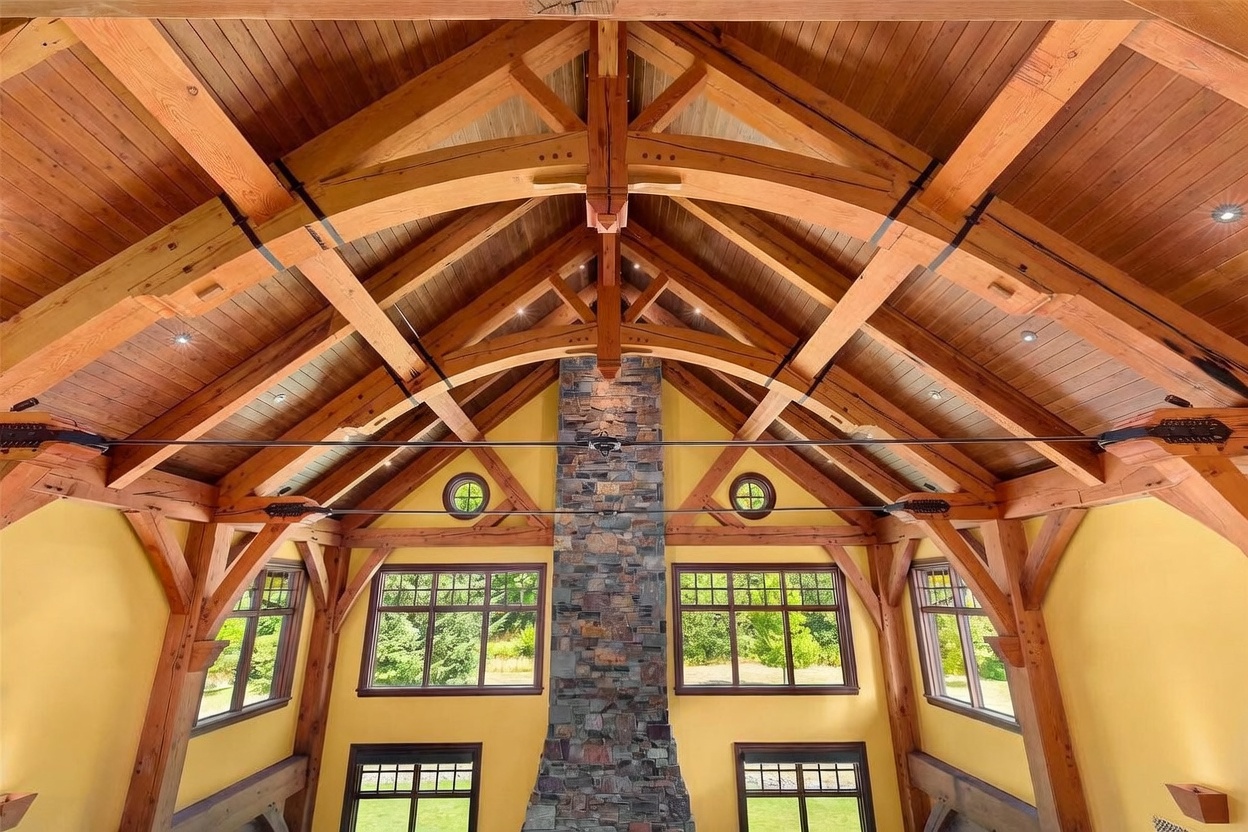 This expansive living area features a dramatic vaulted ceiling with exposed timber trusses and warm wood paneling. A prominent stone-clad chimney breast serves as the central focal point, flanked by large windows that invite natural light into the space. The room is characterized by its rustic, lodge-inspired aesthetic and open, airy atmosphere.