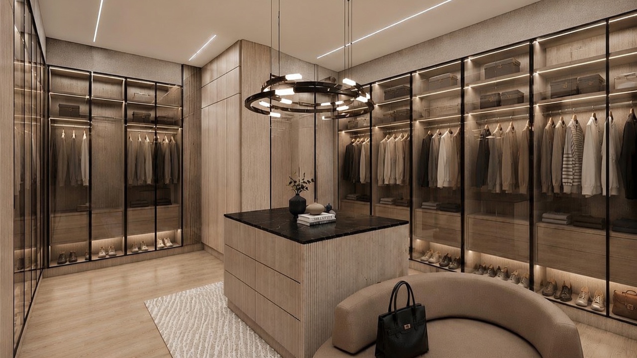 This luxurious walk-in closet features extensive custom cabinetry with glass-front doors and integrated interior lighting. A central island with a dark stone top provides additional storage and surface space, while a curved, upholstered bench offers seating. The room is illuminated by a modern circular chandelier, creating a sophisticated and organized boutique-like atmosphere.