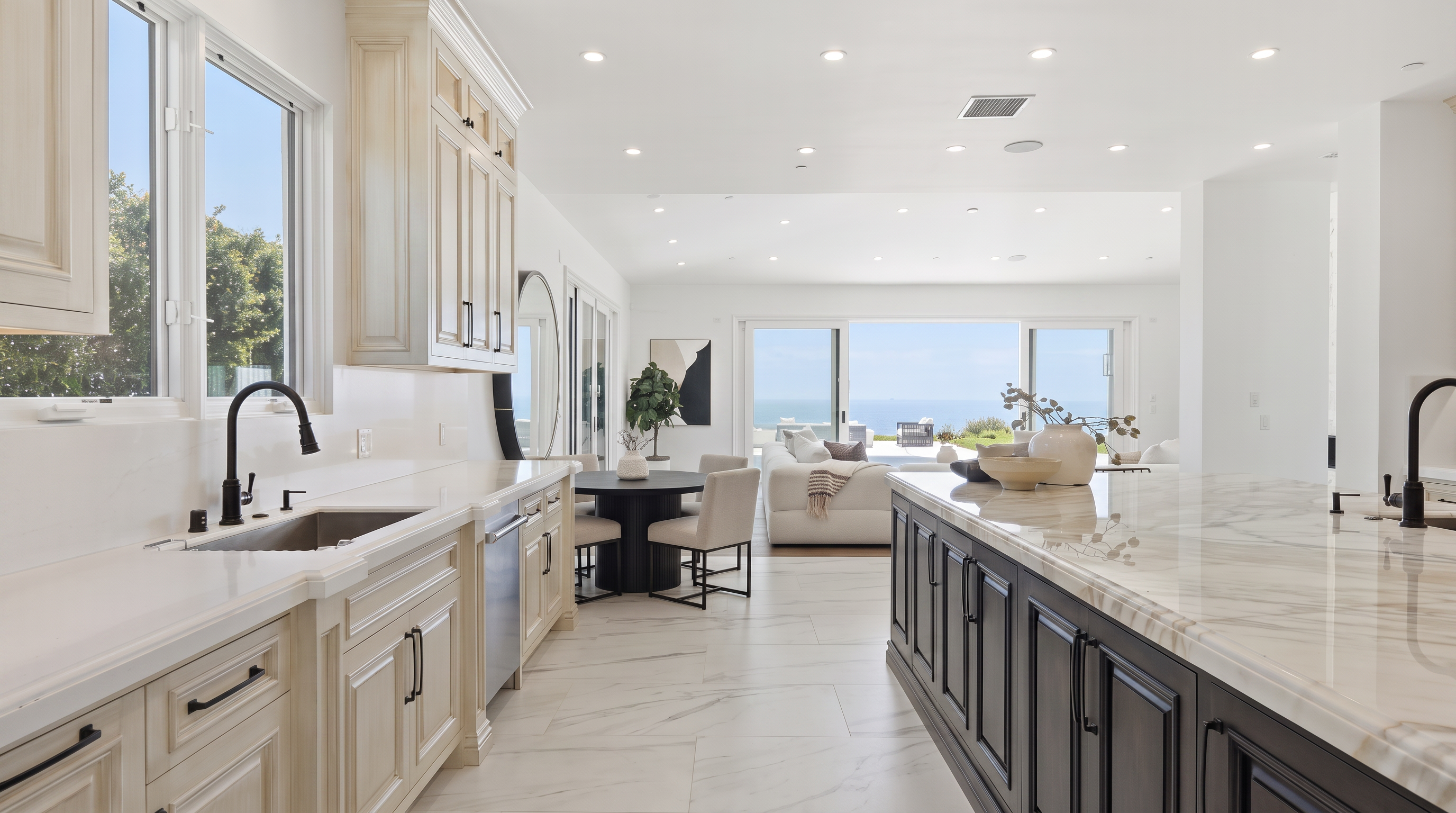 This bright, open-concept kitchen features a large central island with dark cabinetry and a light-toned stone countertop, contrasting with the surrounding light-colored cabinetry. The space flows seamlessly into a dining area and a living room that opens to an expansive ocean view through large sliding glass doors. The neutral color palette and recessed lighting create a clean, airy, and sophisticated atmosphere.