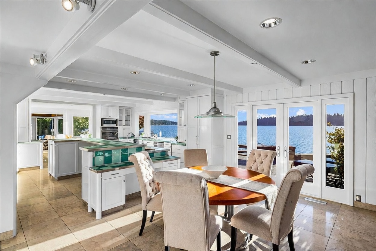 This bright, open-concept kitchen and dining area features a large island with a distinctive green tiled countertop and white cabinetry. A round wooden dining table sits in the foreground, framed by elegant upholstered chairs, while large glass doors offer a stunning, unobstructed view of the lake. The space is illuminated by recessed lighting and a stylish pendant fixture, creating a warm and inviting atmosphere perfect for waterfront living.