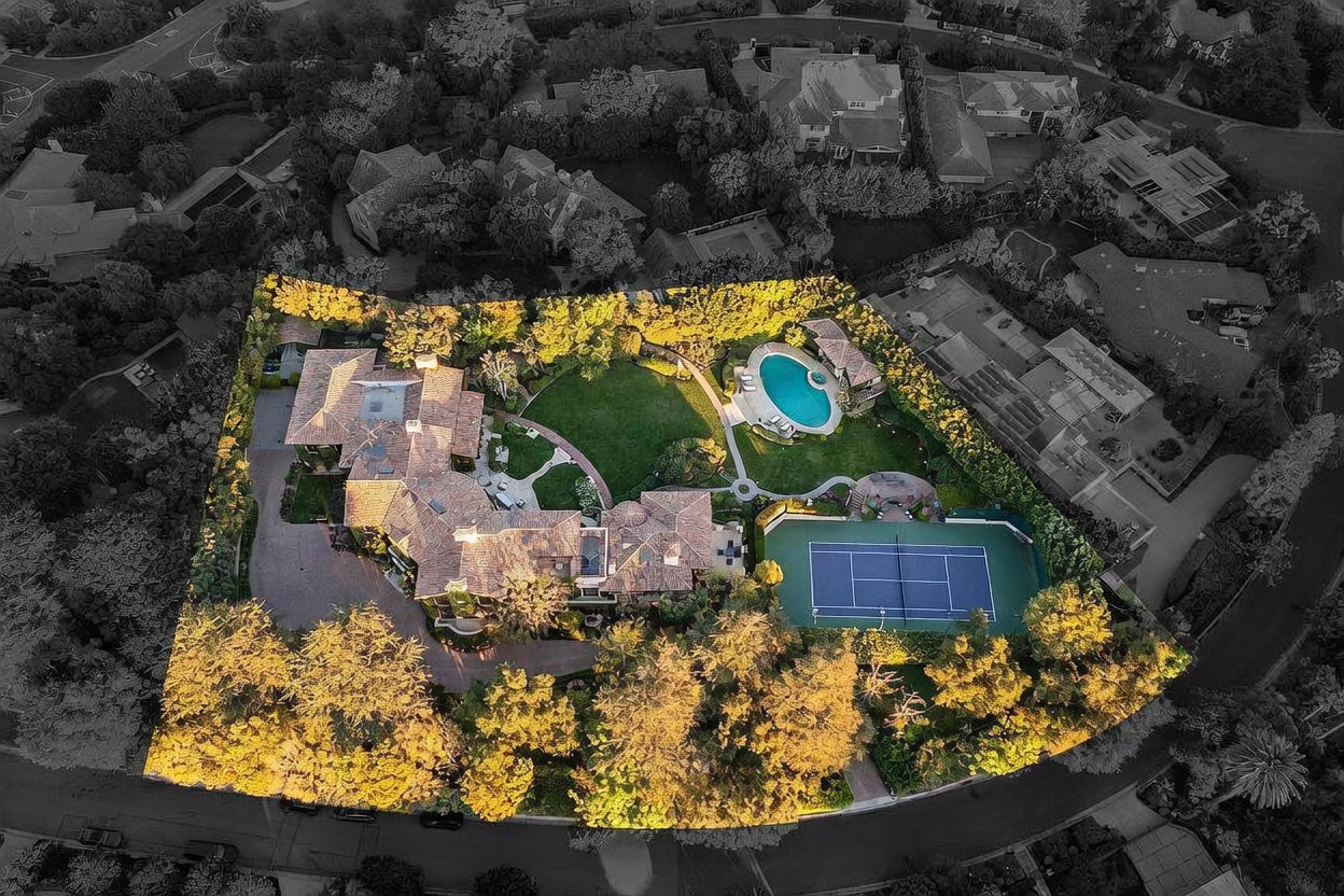This high-angle aerial view captures a sprawling luxury estate nestled within a lush, tree-lined neighborhood. The property features a large multi-winged residence with a tiled roof, a manicured circular lawn, a private swimming pool, and a full-sized tennis court. The perspective highlights the expansive grounds and the secluded, private nature of the estate.