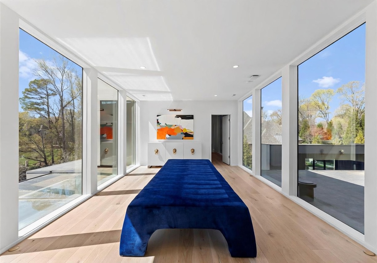 This bright, minimalist room features expansive floor-to-ceiling windows on both sides, offering panoramic views of the surrounding trees and outdoor deck. A long, vibrant blue velvet bench serves as a central focal point, positioned in front of a white console table adorned with a colorful abstract painting. The space is finished with light wood flooring, creating a clean, airy, and modern atmosphere.
