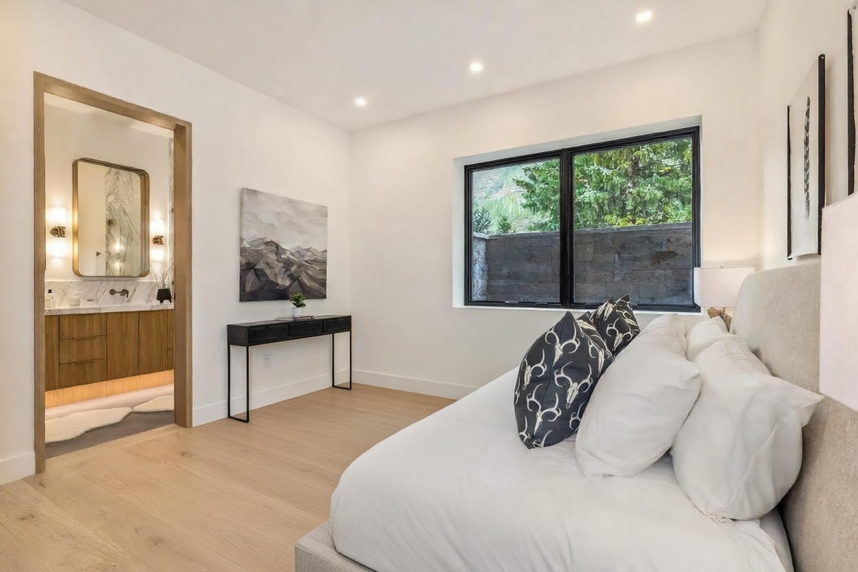 This modern bedroom features a neutral color palette with a plush bed dressed in white linens and a dark gray knit throw. A sleek black console table sits against the wall beneath abstract artwork, while a doorway leads into an adjacent bathroom with a vanity and mirror. Large windows provide natural light and a view of the outdoor stone wall, creating a serene and contemporary atmosphere.