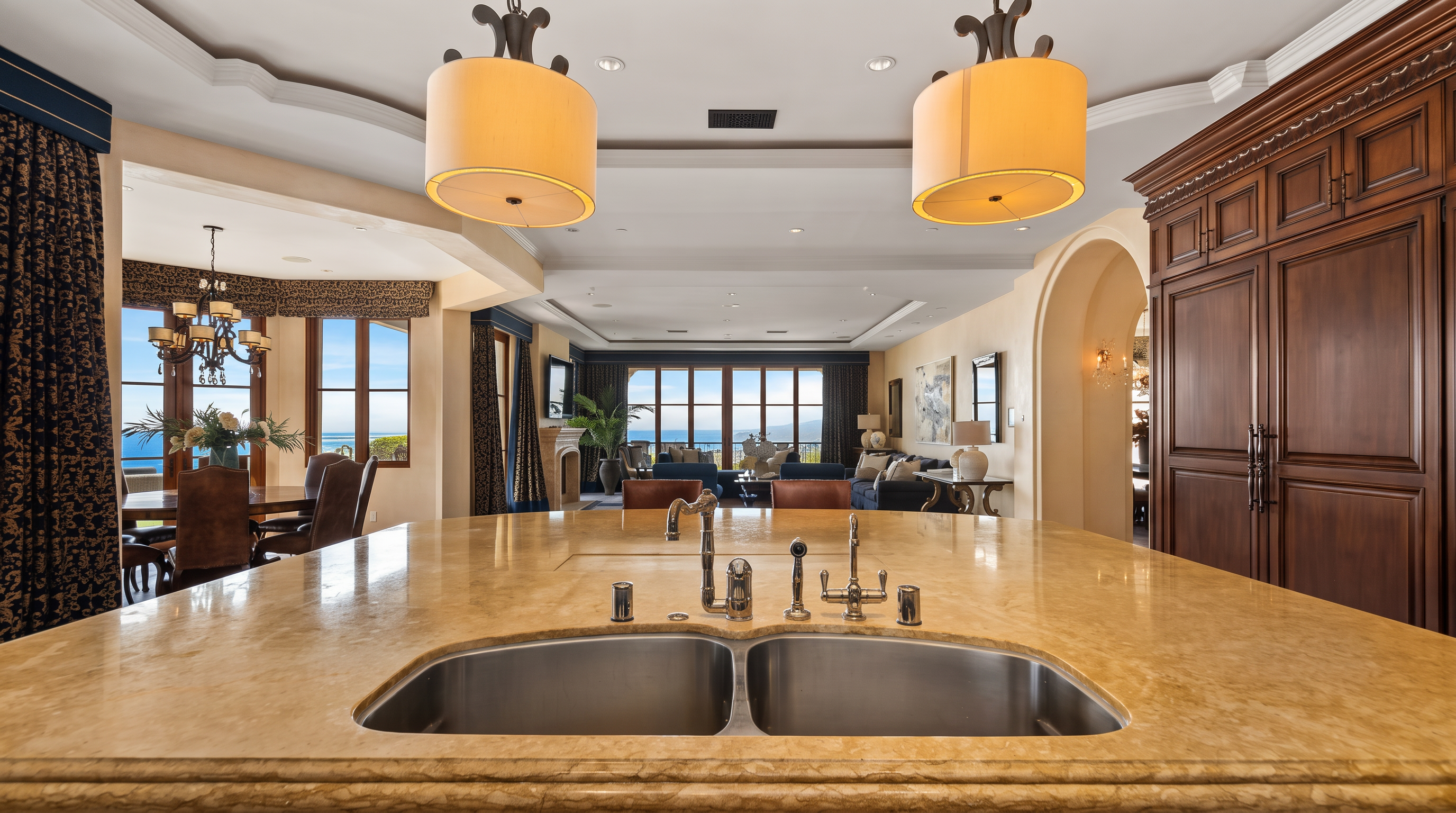 This perspective from the kitchen island looks across an open-concept living area toward a scenic ocean view framed by large windows. The kitchen features a prominent stone-topped island with a double-basin sink and dual pendant lights, while the background reveals a dining area and a comfortable living space. The interior design is characterized by warm tones, traditional cabinetry, and a seamless flow between the culinary and relaxation zones.