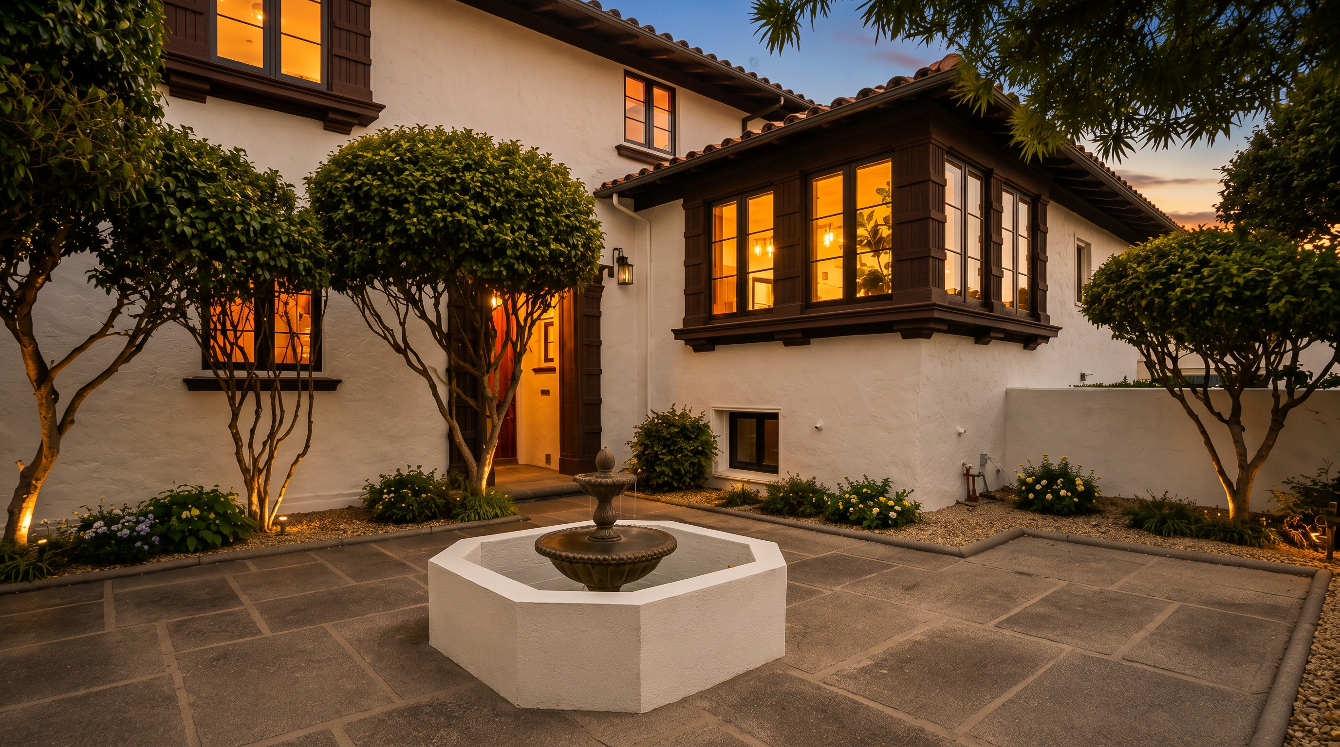 This charming Mediterranean-style home features a private, paved courtyard centered around an elegant stone fountain. The white stucco exterior is accented by dark wood trim and a prominent bay window, creating a warm and inviting entrance. Lush, manicured trees and low-lying shrubs frame the space, enhancing the tranquil and sophisticated atmosphere of the home's front approach.
