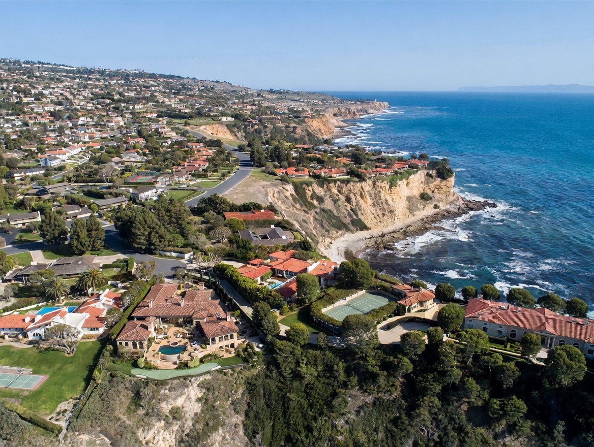 This high-angle aerial perspective captures a sprawling coastal estate perched on a dramatic cliffside overlooking the Pacific Ocean. The property features multiple luxury residences with terracotta-tiled roofs, private swimming pools, and a tennis court, all surrounded by lush landscaping. The expansive view highlights the rugged coastline, winding residential streets, and the vast blue horizon, emphasizing the exclusive and serene nature of this prime waterfront location.