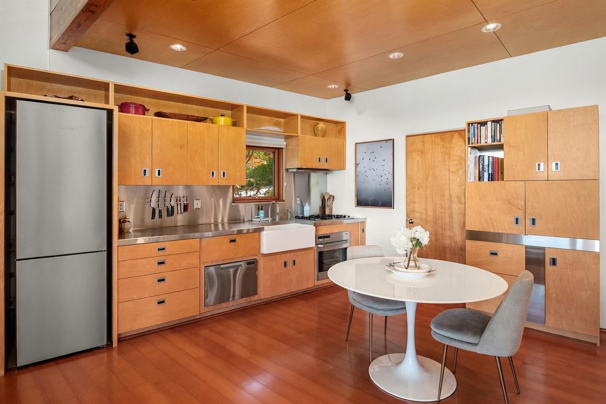 This modern kitchen features light wood cabinetry, stainless steel appliances, and a farmhouse-style sink set into a metallic countertop. A round white dining table with two grey chairs sits in the foreground, while a wood-paneled ceiling adds warmth to the space. The room is well-lit with recessed lighting and includes a small window above the sink, creating a bright and functional culinary area.