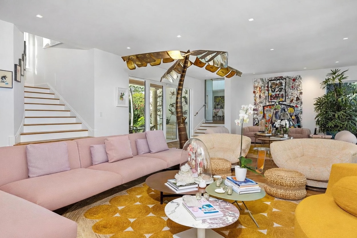 This spacious, brightly lit living room features a large, soft pink sectional sofa paired with textured, cream-colored armchairs and a vibrant yellow circular rug. A unique, sculptural metal palm tree lamp serves as a central focal point, while a large, colorful abstract painting adds artistic flair to the wall. The room is accented by natural textures like woven ottomans and a variety of coffee tables, creating a sophisticated yet playful atmosphere.
