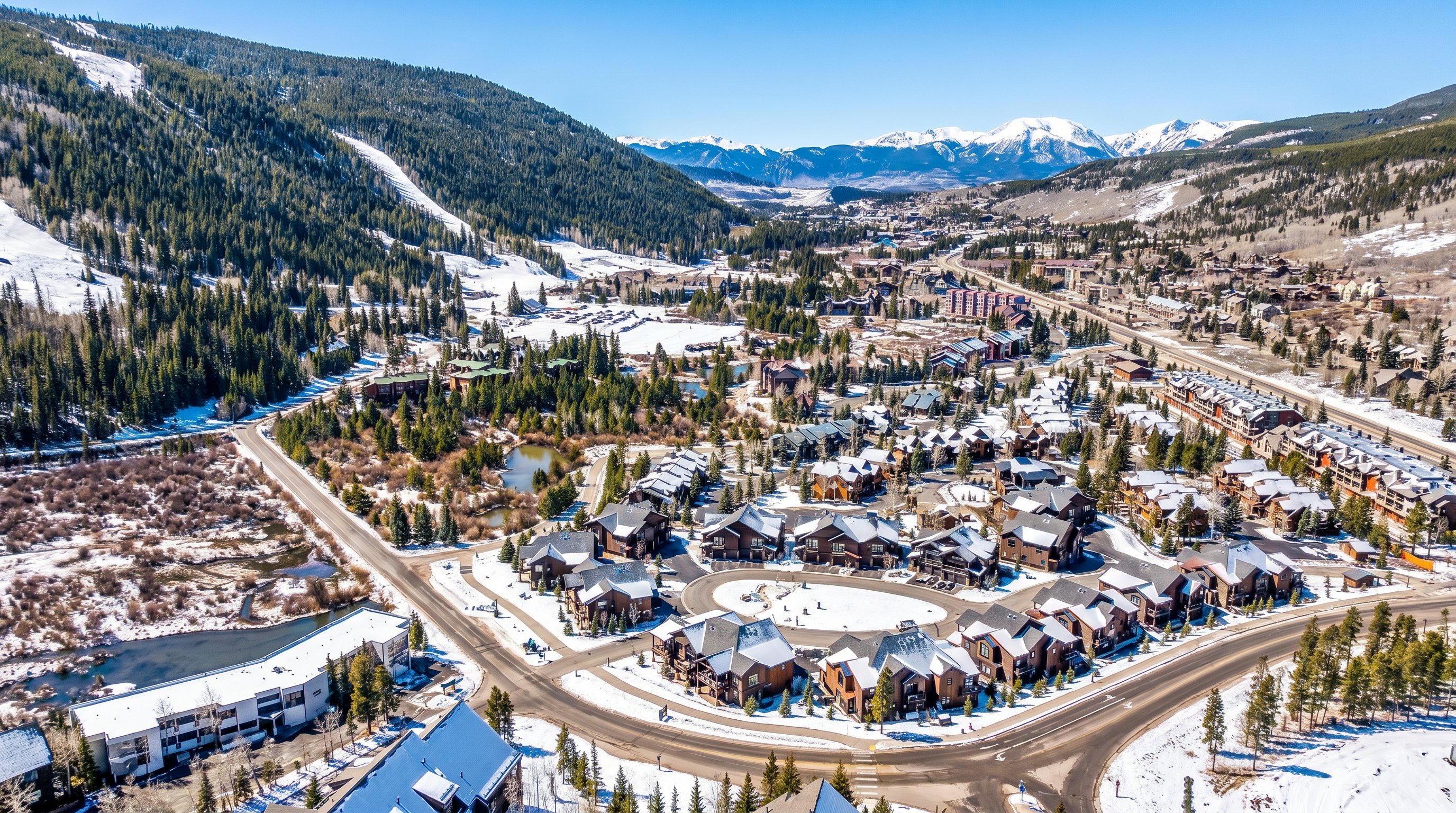 This high-angle aerial perspective captures a sprawling mountain resort community nestled in a lush, forested valley. The scene features a cluster of multi-story residential buildings with dark shingled roofs, winding roads, and a scenic backdrop of snow-capped peaks under a vibrant blue sky. The composition emphasizes the expansive natural landscape and the integrated design of the neighborhood within the alpine environment.