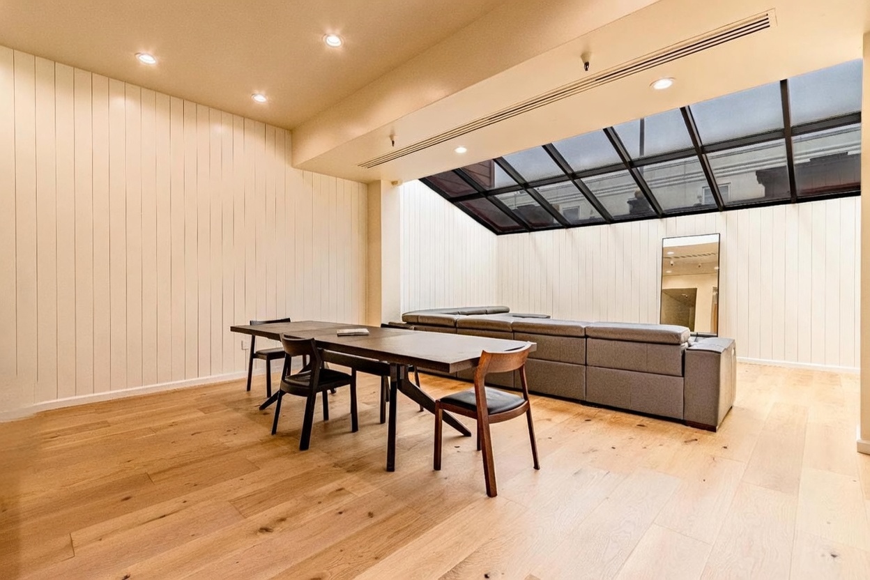 This bright, open-concept living and dining area features light wood flooring and vertical wood paneling on the walls. A large, angled skylight spans the ceiling above a spacious grey sectional sofa, while a dark wood dining table with matching chairs sits in the foreground. The room has a minimalist, modern aesthetic with a tall mirror leaning against the back wall, creating a sense of depth and openness.
