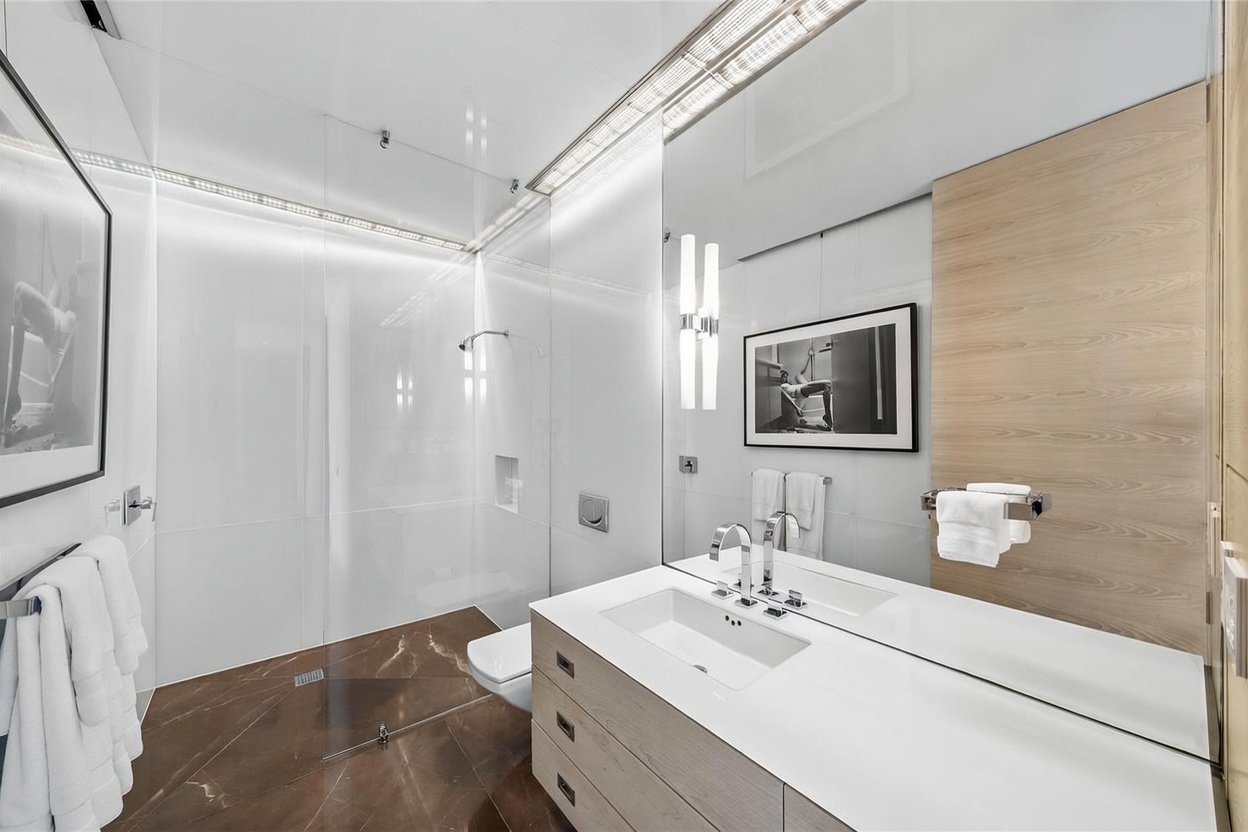 This modern primary bathroom features a sleek, floating light wood vanity with a white countertop and dual faucets. A large, frameless glass-enclosed shower area with dark, patterned stone flooring creates a sophisticated contrast against the bright white walls and ceiling. The space is illuminated by integrated lighting and includes framed artwork, offering a clean, minimalist, and high-end aesthetic.